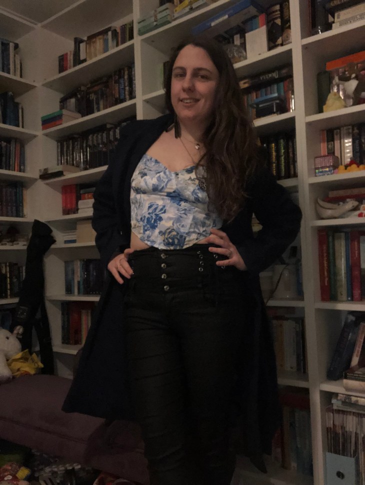 Me dressed for a poetry event in a blue and white corseted crop top, black leather effect trousers with silver buttons, a pair of black heels, and a blue three-quarter length military style coat with my hair loose, and black feather earrings. I'm stood in front of my bookcases at home. 